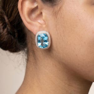 May include: A large, cushion-cut light blue gemstone earring set in a silver-toned metal frame. The central blue stone is surrounded by a halo of small, clear, sparkling stones that line the entire rectangular border of the earring piece.