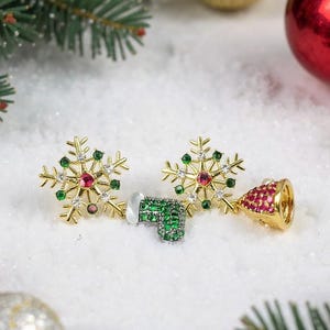 May include: Gold-toned snowflake earrings with red and green gemstones, and a bell-shaped earring with red gemstones. The earrings are set against a snowy background with Christmas tree branches and ornaments.