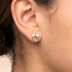 May include: A gold-toned stud earring shaped like a bow, featuring two clear, round-cut stones set at the centre of the loops. The metal frame forms the outline of a ribbon, with the stones positioned to create a sparkling effect on the earlobe.