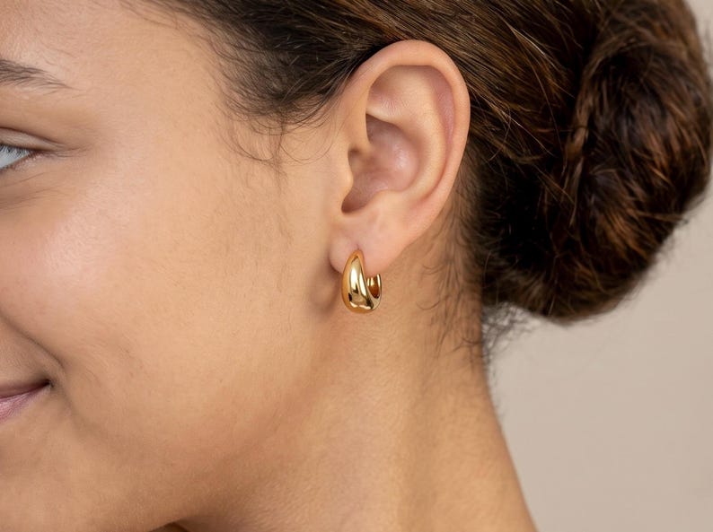 Gold-colored hoop earrings,  10mm in size, are displayed on a left ear. The earrings have a smooth, polished finish and a classic, rounded shape. The image is a close-up, highlighting the jewelry's design and shine.