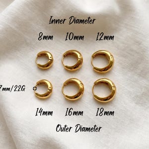 Gold-colored hoop earrings, 8mm, 10mm 12mm inner diameter size with their outer diameter being 15, 16, and 18mm respectively, are displayed on a soft ivory cream fabric. The image is a close-up, highlighting the jewelry's design and shine. Earring post diameter is 0.7mm/22G