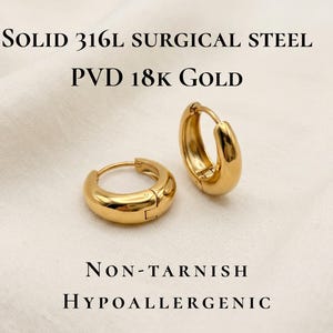 Gold-colored hoop earrings, 12 in size, are displayed on a soft ivory cream fabric with infographic that says they are solid 316l surgical steel with PVD 18k gold, NonTarnish and hypoallergenic. The earrings have a smooth, polished finish and a classic, rounded shape. The image is a close-up, highlighting the jewelry's design and shine.