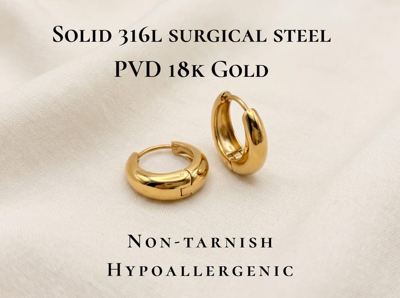 Gold-colored hoop earrings, 12 in size, are displayed on a soft ivory cream fabric with infographic that says they are solid 316l surgical steel with PVD 18k gold, NonTarnish and hypoallergenic. The earrings have a smooth, polished finish and a classic, rounded shape. The image is a close-up, highlighting the jewelry's design and shine.