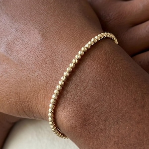 May include: A gold beaded bracelet. The bracelet is made of small, round beads strung together to form a flexible band. The bracelet is worn on a person's wrist, and the gold color gives it a classic look.