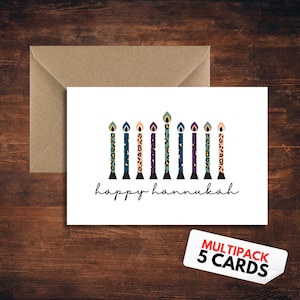 Könnte beinhalten: Weiße Karte mit einem Menora-Design und dem Schriftzug "happy hannukah" in schwarzer Schrift. Die Menora-Kerzen haben farbenfrohe Leopardenmuster. Enthält einen Kraftpapierumschlag und einen Aufkleber mit der Aufschrift "MULTIPACK 5 CARDS".