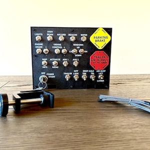 Puede incluir: Panel de control negro con múltiples interruptores para luces, motor y funciones del remolque. Incluye una llave, una abrazadera y un cable. El panel tiene las etiquetas "FRENO DE ESTACIONAMIENTO" y "SUMINISTRO DE AIRE DEL REMOLQUE".