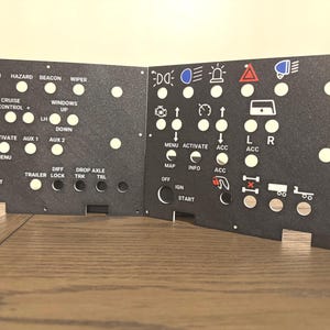 May include: Black control panel with white text and icons. Features buttons labeled "Lights", "High Beam", "Hazard", "Beacon", "Wiper", "Engine Brake", "Cruise Control", and "Windows Up". Includes icons for lights, arrows, and a trailer.