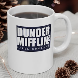 May include: White ceramic mug with a blue and white logo that says "Dunder Mifflin, Inc. Paper Company". The mug is filled with coffee and steam is rising from the top.