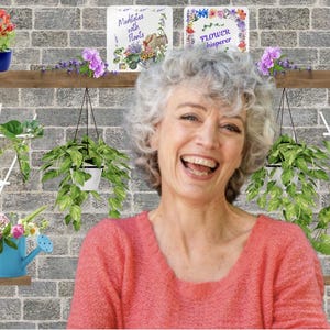 May include: A woman with gray hair smiles while standing in front of a brick wall with hanging plants and two books titled "Meditates with Plants" and "Flower Whisperer".