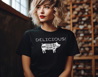 Könnte beinhalten: Ein schwarzes T-Shirt mit einer weißen Grafik eines Schweins mit verschiedenen Fleischstücken, die beschriftet sind. Der Text "DELICIOUS!" befindet sich über dem Schwein.