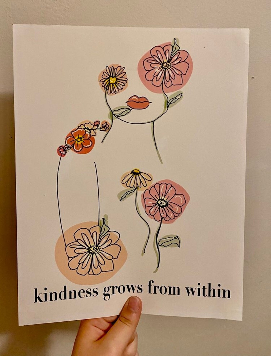 May include: A white background with a line drawing of a face with flowers in place of the eyes and a flower on the shoulder. The text 'kindness grows from within' is at the bottom of the image.