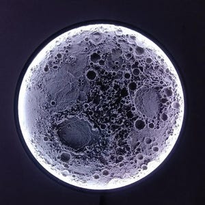 Op de afbeelding: Een ronde, verlichte maanlamp met een gedetailleerde oppervlaktestructuur, met kraters en verschillende grijstinten. De lamp heeft een heldere, witte buitenring, waardoor een halo-effect ontstaat tegen een donkere achtergrond. Het is een decoratief item.