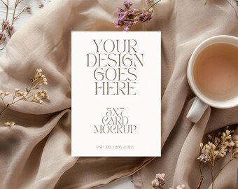 May include: A white rectangular card mockup with the brown text YOUR DESIGN GOES HERE 5X7 CARD MOCKUP PSP JPG CANVA FILE, placed on beige fabric next to a white ceramic mug and small sprigs of dried pink and brown flowers.