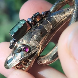 May include: A gold-toned snake ring with a detailed, textured body. The snake's head features a ruby eye and is adorned with green and silver-toned gemstones. The ring is held in a hand, showcasing the intricate design and craftsmanship.