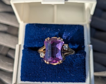 Può includere: Un anello in oro con una grande pietra preziosa rettangolare sfaccettata di colore viola al centro, affiancata da due piccole pietre rotonde e chiare su ciascun lato, presentato all'interno di un portagioie bianco con interno in velluto blu.