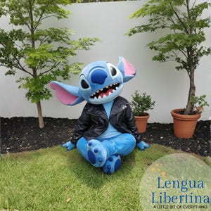 May include: A blue Stitch costume with large ears and a black leather jacket is seated on the grass. The costume has large eyes and a wide grin. The costume is in a garden setting with small trees and the text "Lengua Libertina" in the bottom right corner.
