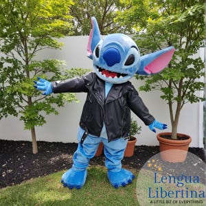 May include: A blue character costume with large ears, wearing a black leather jacket and blue pants. The character has a wide smile and is standing in front of green trees. The costume has blue hands and feet.