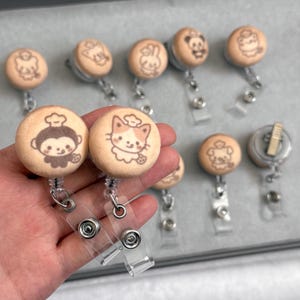 May include: A collection of round badge reels featuring light tan fabric covers with laser-engraved illustrations of various cartoon animals wearing chef hats. Each reel includes a clear plastic strap and a metal clip for attachment.