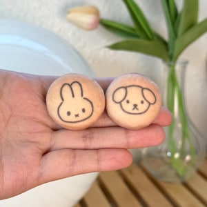 May include: Two small, round, beige-colored objects with black line drawings of a bunny and a puppy. The bunny has long ears and a simple face, while the puppy has floppy ears and a small nose. The objects are held in a hand.