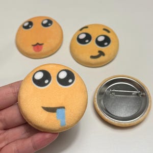 Könnte beinhalten: Vier runde, orangefarbene Emoji-Buttons mit verschiedenen Ausdrücken. Ein Button hat eine herausgestreckte Zunge, ein anderer ein lächelndes Gesicht und ein dritter einen sabbernden Ausdruck. Der vierte Button zeigt die Rückseite mit einer Nadel.