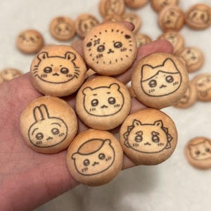 Könnte beinhalten: Kleine, runde Kekse mit verschiedenen Cartoon-Tiergesichtern, die mit schwarzer Tinte gezeichnet wurden. Zu den Designs gehören ein Bär, eine Katze und ein Hase. Die Kekse sind hellbraun.