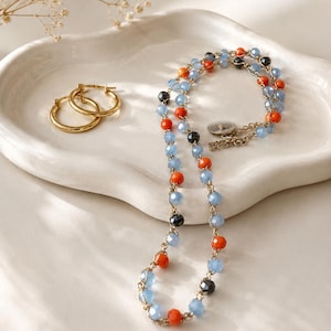 May include: A beaded necklace featuring light blue, orange, and black faceted beads strung on a gold-toned chain with a circular clasp, accompanied by a pair of simple gold-toned hoop earrings. The jewelry set is arranged on a white ceramic tray.