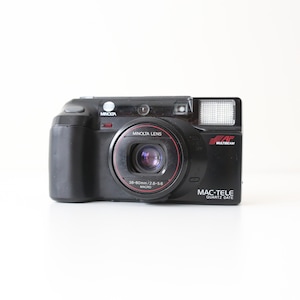 May include: Black Minolta Mac-Tele Quartz Date camera with a red-ringed lens. The camera has a built-in flash and the Minolta Lens is 38-80mm/2.8-5.6 Macro. The camera has the text "AF Multibeam" and "Minolta" printed on it.
