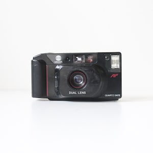 May include: A black Minolta AF Quartz Date dual lens camera. The camera has a red accent on the side and the words "DUAL LENS" and "QUARTZ DATE" are printed on the front. The camera has a flash and a lens.