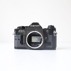 Könnte beinhalten: Schwarze Canon AE-1 Program Kamera ohne Objektiv. Das Kameragehäuse trägt das Canon-Logo und den Text AE-1 Program. Die Kamera hat silberne Metallakzente und einen strukturierten Griff.