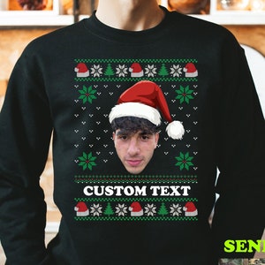 Op de afbeelding: Een zwart sweatshirt met lange mouwen en een feestelijk patroon van groene sneeuwvlokken en rode kerstmutsen. In het midden staat een aangepast portret van een persoon met een kerstmuts boven de witte tekst CUSTOM TEXT.