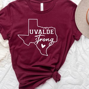 May include: Burgundy t-shirt with a white outline of the state of Texas and the words "UVALDE Strong" in a script font. A small white heart is inside the outline. The shirt is knotted at the bottom.