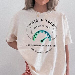 May include: A cream-colored short-sleeve t-shirt featuring a graphic of a speedometer with the text THIS IS YOUR BADDNESS LEVEL IT'S UNSUSUALLY HIGH FOR SOMEONE YOUR SIZE and an outline of two ears, paired with red and white gingham shorts.
