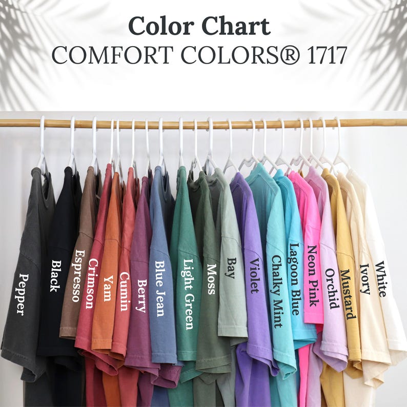 May include: A color chart displays a variety of Comfort Colors 1717 t-shirts hanging on white hangers. The shirts are in various colors, including black, crimson, blue jean, and neon pink. The text "Color Chart" and "COMFORT COLORS&reg; 1717" are at the top.