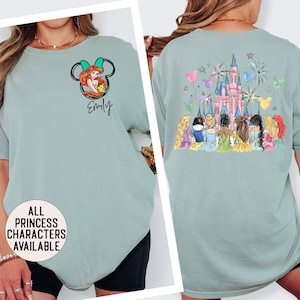 May include: A light green t-shirt featuring a cartoon image of Ariel from The Little Mermaid inside a Mickey Mouse head. The name "Emily" is written below the image. The back of the shirt shows a pink castle, fireworks, and Disney princesses.