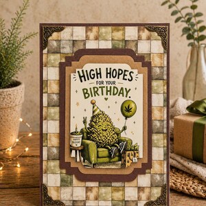 May include: A birthday card with a cartoon illustration. The card features a cannabis bud character wearing a party hat, holding a cannabis leaf balloon, and sitting in a green armchair. The text reads "High Hopes for your Birthday."