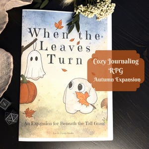 Op de afbeelding: Een softcover boek getiteld When the Leaves Turn, een uitbreiding voor Beneath the Tall Grass. De kaft bevat een aquarelillustratie van een geest, een pompoen en vallende oranje bladeren. Tekst: Cozy Journaling RPG Autumn Expansion.