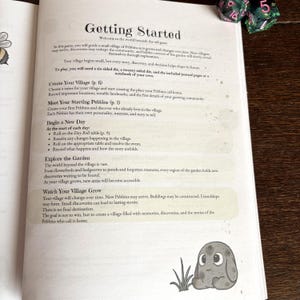 May include: A page from a game book titled Getting Started, featuring instructions for a village simulation game. The text outlines steps for creating a village, meeting characters, and exploring a garden. A small gray creature illustration sits at the bottom.