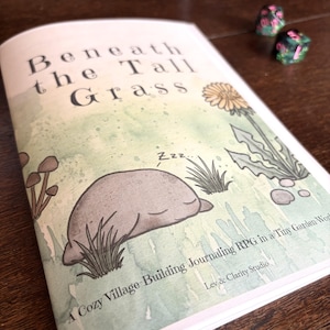 May include: A softcover book titled Beneath the Tall Grass with a watercolor illustration of a sleeping creature, mushrooms, and a dandelion. The cover text reads A Cozy Village-Building Journaling RPG in a Tiny Garden World by Lev & Clarity Studio.