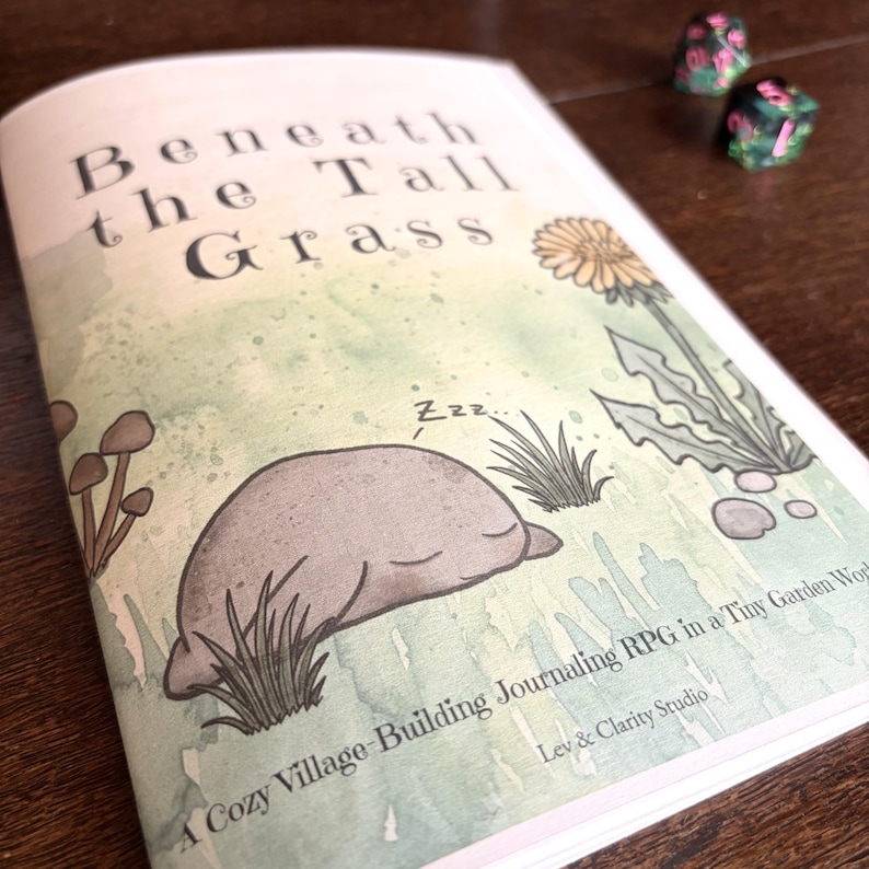 May include: A softcover book titled Beneath the Tall Grass with a watercolor illustration of a sleeping creature, mushrooms, and a dandelion. The cover text reads A Cozy Village-Building Journaling RPG in a Tiny Garden World by Lev & Clarity Studio.