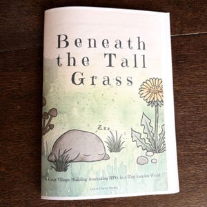 May include: A softcover book titled Beneath the Tall Grass with a light green watercolor background. The cover features a sleeping gray rock, a yellow dandelion, and small mushrooms. Text reads: A Cozy Village-Building Journaling RPG in a Tiny Garden World, Lev & Clarity Studio.