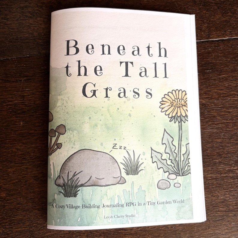 May include: A softcover book titled Beneath the Tall Grass with a light green watercolor background. The cover features a sleeping gray rock, a yellow dandelion, and small mushrooms. Text reads: A Cozy Village-Building Journaling RPG in a Tiny Garden World, Lev & Clarity Studio.