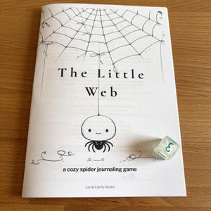 May include: A white booklet titled "The Little Web" with a spider web illustration and a cute spider character. A translucent, green-tinted die with the number 3 is next to the booklet. The text "a cozy spider journaling game" is also visible.