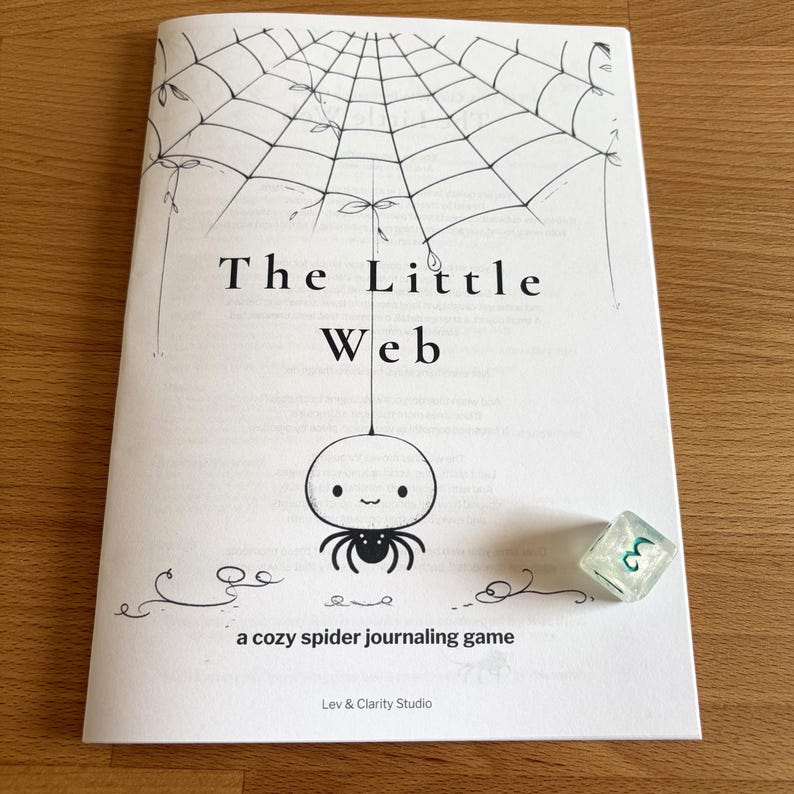 May include: A white booklet titled "The Little Web" with a spider web illustration and a cute spider character. A translucent, green-tinted die with the number 3 is next to the booklet. The text "a cozy spider journaling game" is also visible.