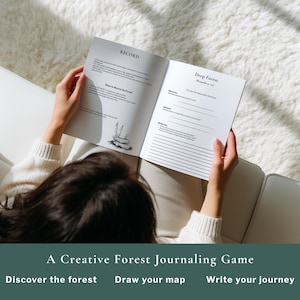May include: A person holding open a journal with the text "RECORD" and "Deep Forest" visible. The journal is part of "A Creative Forest Journaling Game". The person is sitting on a white couch with a fluffy white blanket.