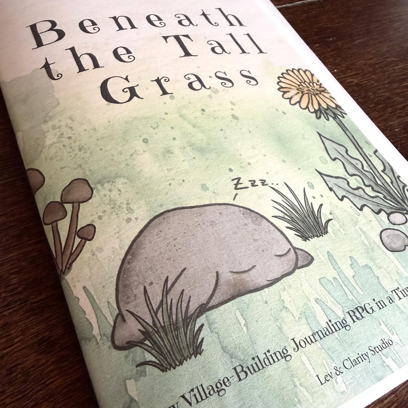 May include: A softcover book titled Beneath the Tall Grass with a watercolor illustration of a sleeping gray creature, brown mushrooms, and a yellow dandelion. The cover features text reading Beneath the Tall Grass and Lev & Clarity Studio.