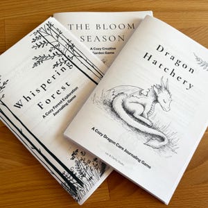 Könnte beinhalten: Drei Broschüren mit Schwarz-Weiß-Illustrationen und Text. Die Titel lauten "Whispering Forest", "Dragon Hatchery" und "The Bloom Season". Das Buch "Dragon Hatchery" zeigt eine Zeichnung eines schlafenden Drachen. Die Bücher liegen auf einer Holzoberfläche.