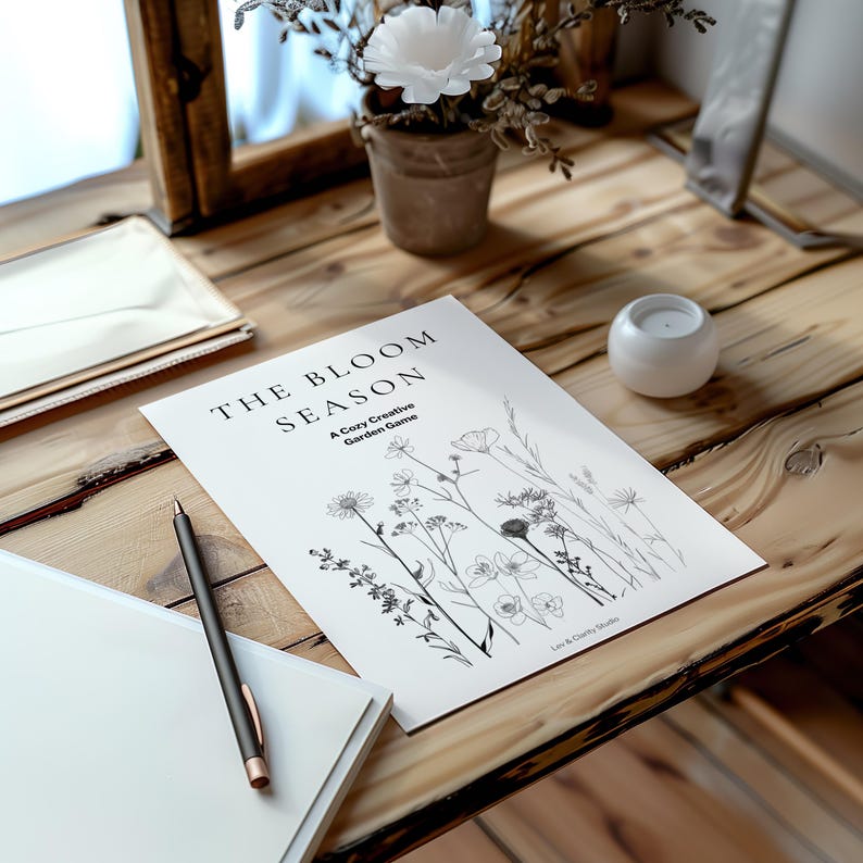 May include: A white paper with the text "THE BLOOM SEASON" and a floral illustration, along with a pen and a notebook, sits on a wooden table. A small potted plant and a candle holder are also on the table.