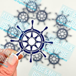 May include: A close-up of a blue and white embroidered iron-on patch with a nautical wheel design. The patch is surrounded by other patches with the text "Aloe & Milk Boutique" on them.