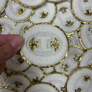 May include: A pile of white felt patches with a gold glitter trim. Each patch has a gold glitter letter "T" in the center.