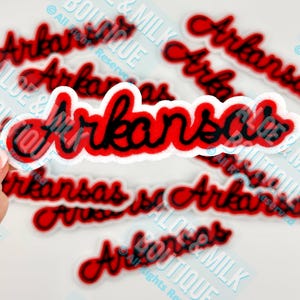 May include: Red and black embroidered patches with the word "Arkansas" in a cursive font. The patches have a white border. The image shows multiple patches, with one held by a hand.
